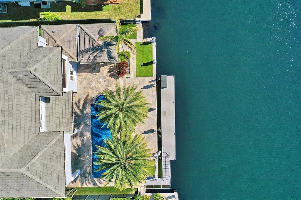 This aerial shot showcases a luxurious waterfront property featuring a large house with a gray roof, a swimming pool surrounded by palm trees, and a private dock extending into the water. The outdoor area includes a patio with a pergola, manicured lawns, and lush landscaping, creating a serene and upscale living environment. The turquoise water adds to the property's appeal, highlighting its prime location.