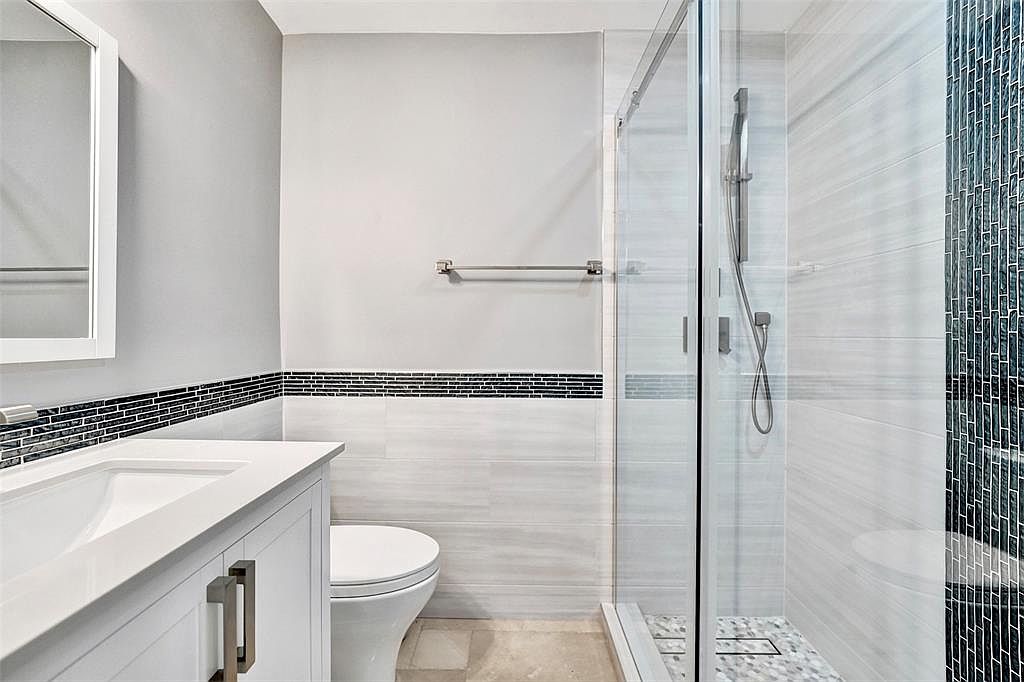 This is a well-lit bathroom featuring a white vanity with a modern sink and chrome hardware, a toilet, and a glass-enclosed shower with tiled walls. A decorative tile border adds a touch of elegance, and the overall design is clean and contemporary. The perspective is from the doorway, showcasing the layout and key features.