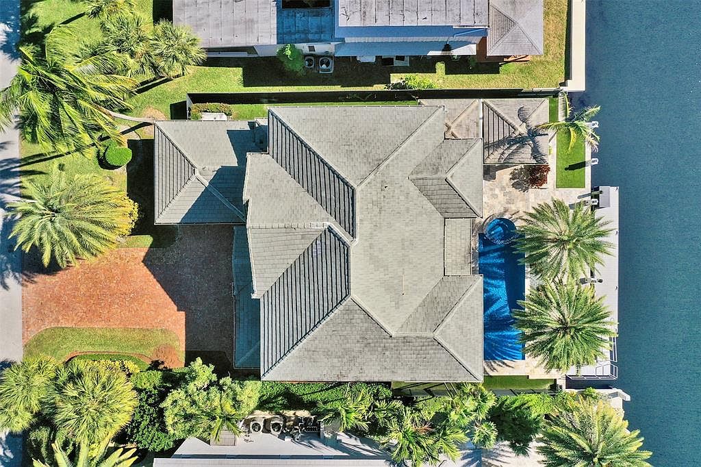 This aerial shot showcases a luxurious home with a complex roofline, a private pool, and direct waterfront access. The property is surrounded by lush landscaping, including mature palm trees, and features a brick driveway. The overall impression is one of high-end coastal living.