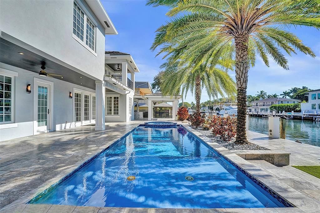 This is a stunning rear exterior view showcasing a luxurious pool and spa area. The rectangular pool features a vibrant blue tile interior, reflecting the sky and surrounding palm trees. The property backs onto a waterfront canal, adding to the upscale and serene ambiance, perfect for relaxation and entertaining.