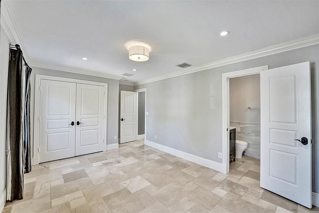 This is a spacious primary bedroom featuring neutral gray walls, white trim, and light-colored tile flooring. The room includes a double-door closet, a doorway leading to another room, and an open bathroom entrance. The overall impression is clean and bright, offering a serene and comfortable atmosphere.