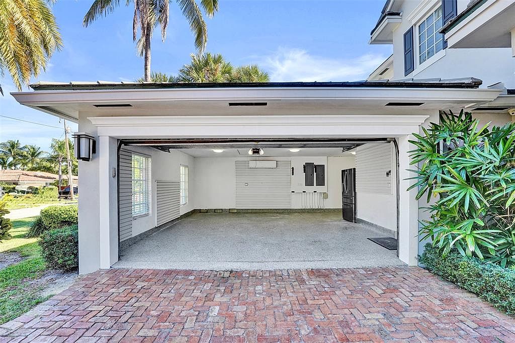 This image showcases an open garage with a brick driveway leading into it. The garage interior is well-lit and appears to be freshly painted, featuring a clean, organized space. The exterior is framed by lush greenery and palm trees, adding to the property's curb appeal.