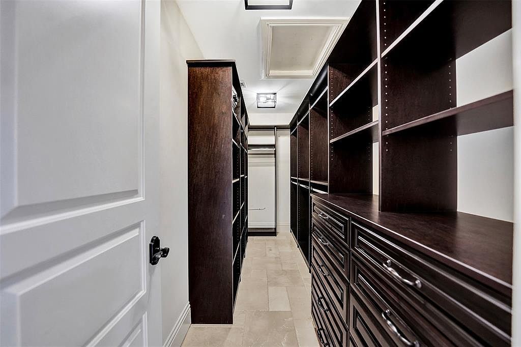 This is a well-organized walk-in closet featuring dark wood shelving and drawers on both sides of a narrow walkway. A white door is partially visible on the left, and the closet is illuminated by overhead lighting. The overall impression is one of luxury and efficient storage.