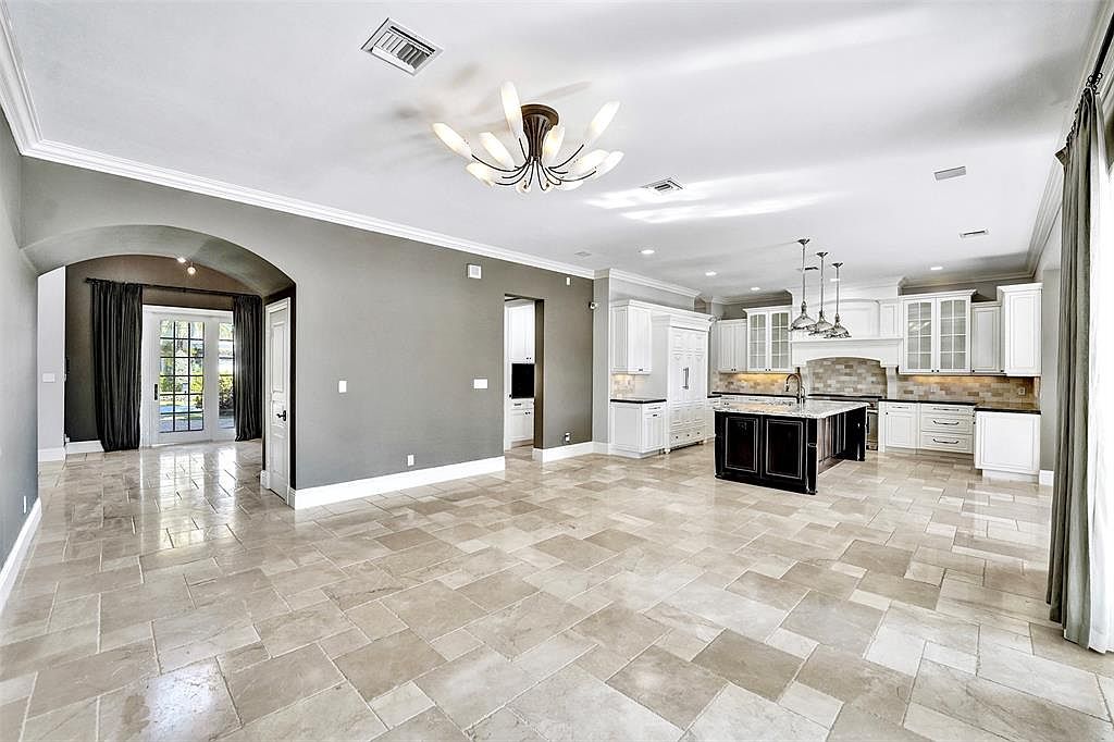 This is a spacious, open-concept kitchen and living area featuring light-colored tile flooring and a neutral color palette. The kitchen boasts white cabinetry, a dark island with a light countertop, and stainless steel appliances, while the living area includes an arched doorway and large windows with dark curtains. The overall impression is one of elegance and modern comfort.