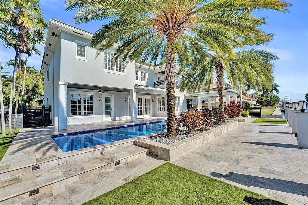 This image showcases the rear exterior of a luxurious home, highlighting a stunning pool and patio area. The pool's clear blue water contrasts beautifully with the light-colored stone patio and lush green lawn. Palm trees add a tropical touch, enhancing the property's appeal and creating a relaxing outdoor oasis.