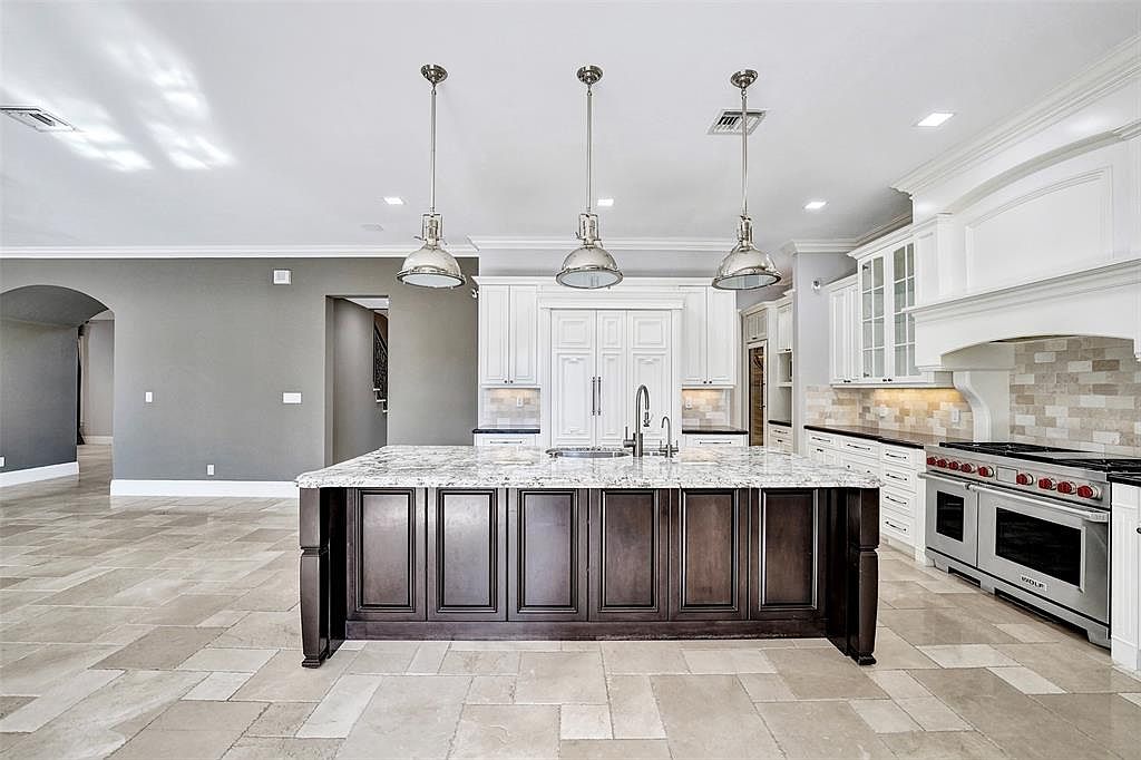 This is a spacious kitchen featuring a large center island with dark wood cabinetry and a light-colored countertop. The kitchen is equipped with stainless steel appliances, including a professional-grade range, and white cabinetry that extends to the ceiling. The flooring is a light-colored tile, and pendant lights hang above the island, creating a bright and inviting atmosphere.