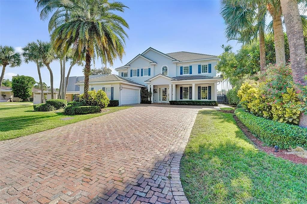 This is a front exterior view of a two-story house with a well-manicured lawn and a brick driveway. The house features a light blue facade, dark shutters, and a covered entryway with columns. Palm trees and other landscaping elements add to the property's curb appeal, creating a welcoming and upscale impression.
