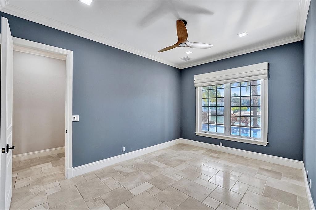 This is a well-lit bedroom with blue walls, white trim, and tile flooring. A window with a roman shade offers a view of the outdoors, possibly a pool or canal. The room also features a ceiling fan and an open doorway leading to another room or closet, creating a sense of spaciousness.