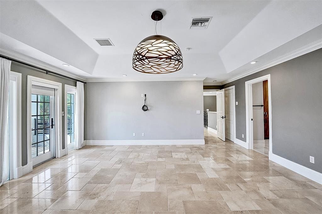 This is a spacious living room featuring travertine tile flooring and neutral gray walls. A modern, decorative pendant light hangs from the white tray ceiling. French doors lead to an outdoor area, and doorways provide access to other parts of the house, creating an open and airy feel.
