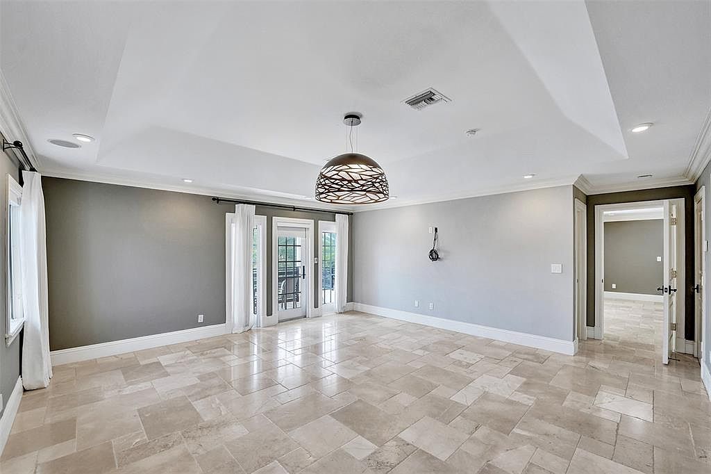 This is a spacious living room featuring neutral-toned walls, light-colored tile flooring, and a modern chandelier. The room has a tray ceiling with recessed lighting and a set of French doors leading to an outdoor area. The overall impression is clean, bright, and inviting, perfect for showcasing the home's open layout.