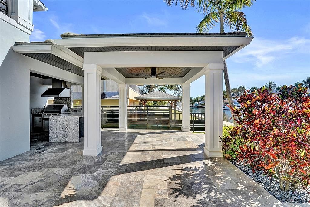 This image showcases a luxurious outdoor patio area with a covered structure supported by white columns. The patio features elegant stone flooring and an outdoor kitchen area with a grill and countertop. Lush landscaping surrounds the patio, adding to the serene and upscale ambiance, with a view of the water in the background.