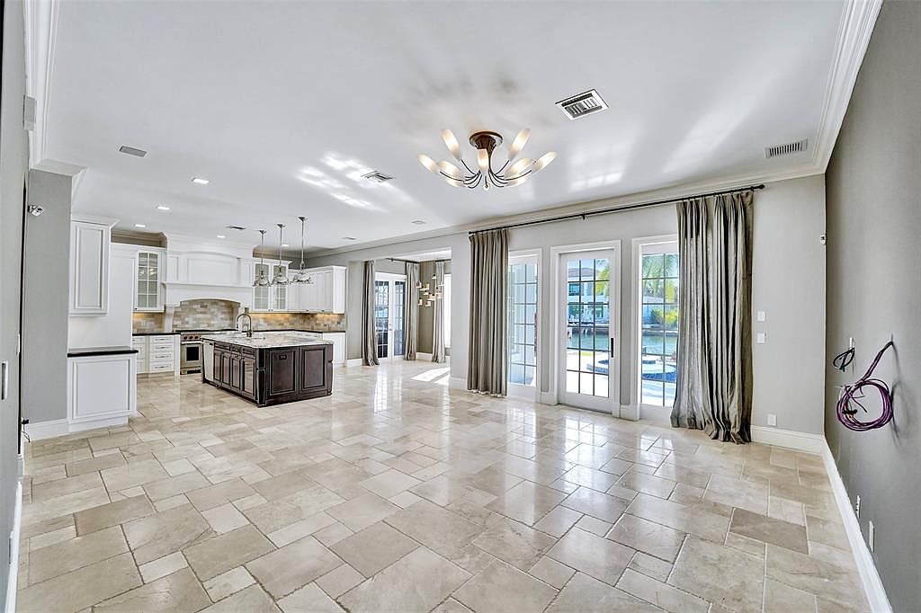 This is a spacious living room that seamlessly connects to the kitchen area. The room features light-colored tile flooring, a modern chandelier, and large windows with curtains that provide natural light and views of the water. The kitchen has white cabinetry and a dark wood island, creating a bright and inviting atmosphere.