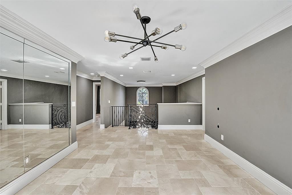 This interior shot showcases a spacious hallway with a modern chandelier and neutral gray walls. The flooring is a light-colored tile arranged in a geometric pattern. A large mirror on the left wall reflects the hallway, enhancing the sense of space, while a staircase with wrought iron railings is visible in the background.