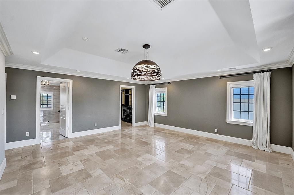 This is a spacious primary bedroom featuring a tray ceiling with recessed lighting and a modern chandelier. The walls are painted in a neutral gray tone, complemented by white trim and baseboards. The flooring is a light-colored tile laid in a geometric pattern, and the room has two windows with white curtains, providing natural light.