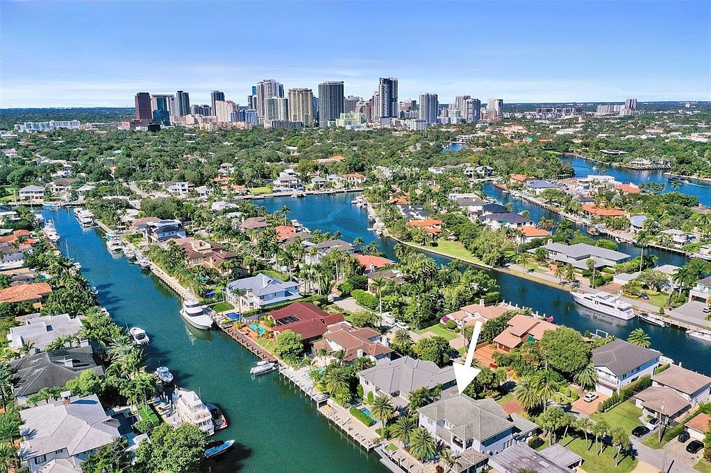 This aerial view showcases a luxurious waterfront property nestled along a canal, complete with a private dock and boat. The house features a gray roof, lush landscaping, and a spacious backyard. In the background, a vibrant cityscape adds to the property's appeal, highlighting its prime location and upscale neighborhood.