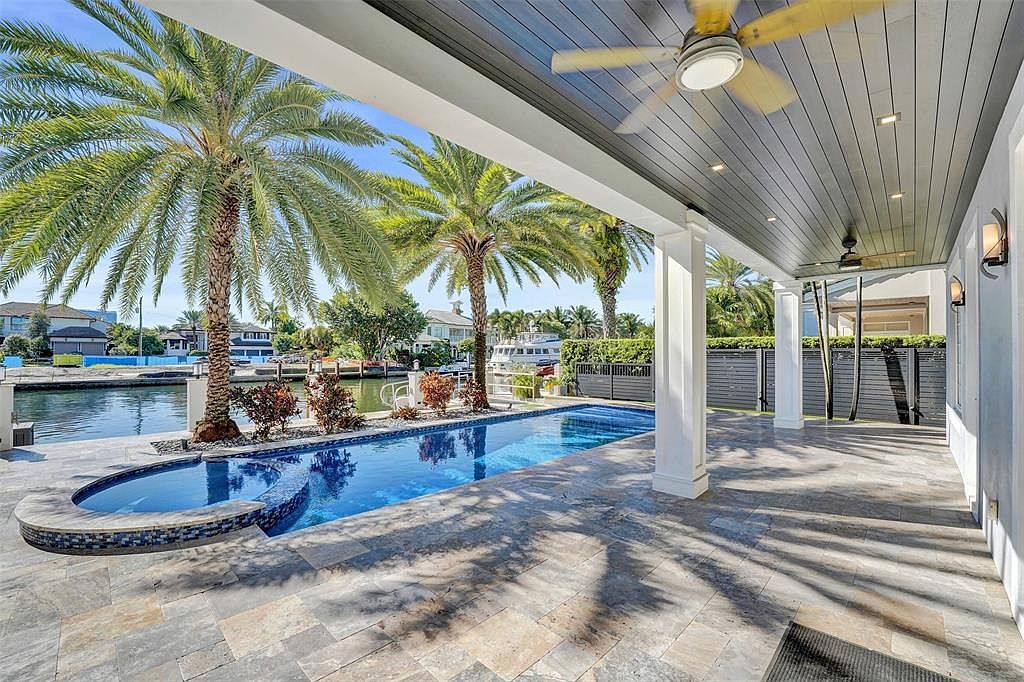 This image showcases a luxurious outdoor pool area with a sleek, modern design. The pool is surrounded by palm trees and a stylish patio with gray stone tiles. A covered patio area with white columns and ceiling fans provides shade and adds to the upscale ambiance, perfect for relaxation and entertainment.