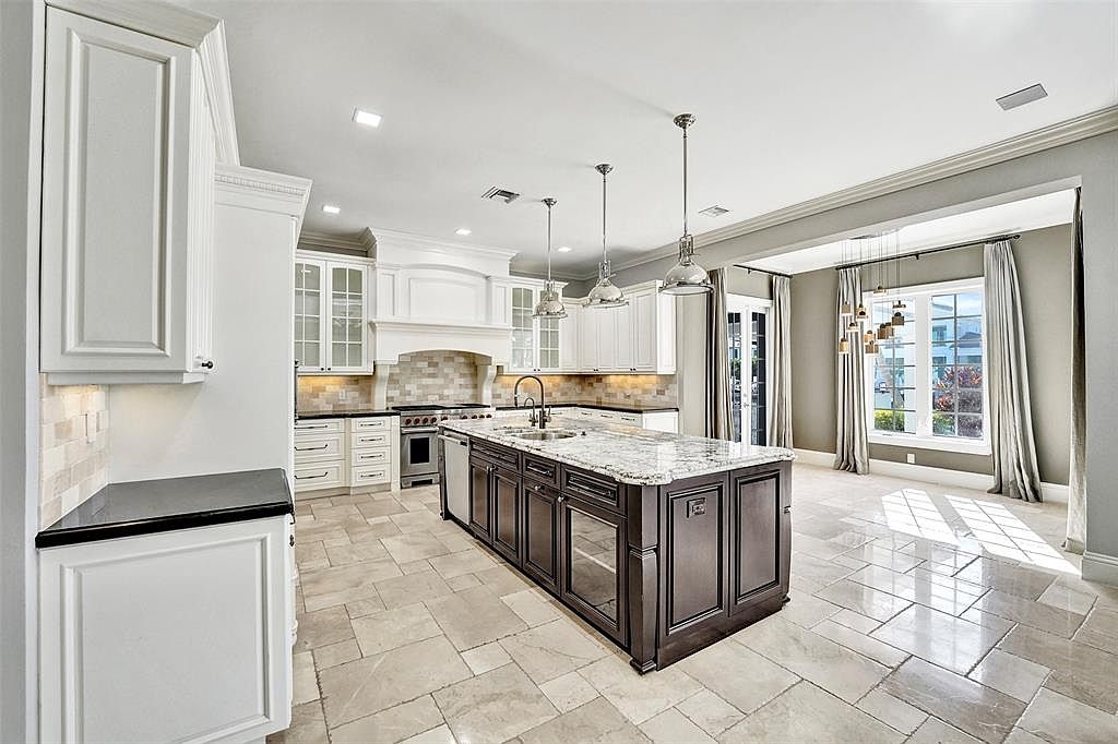 This is a spacious kitchen featuring white cabinetry, a dark wood island with a light countertop, and stainless steel appliances. The kitchen has a classic design with modern lighting fixtures and a tile floor. The perspective is wide, showcasing the entire kitchen and the adjacent dining area.