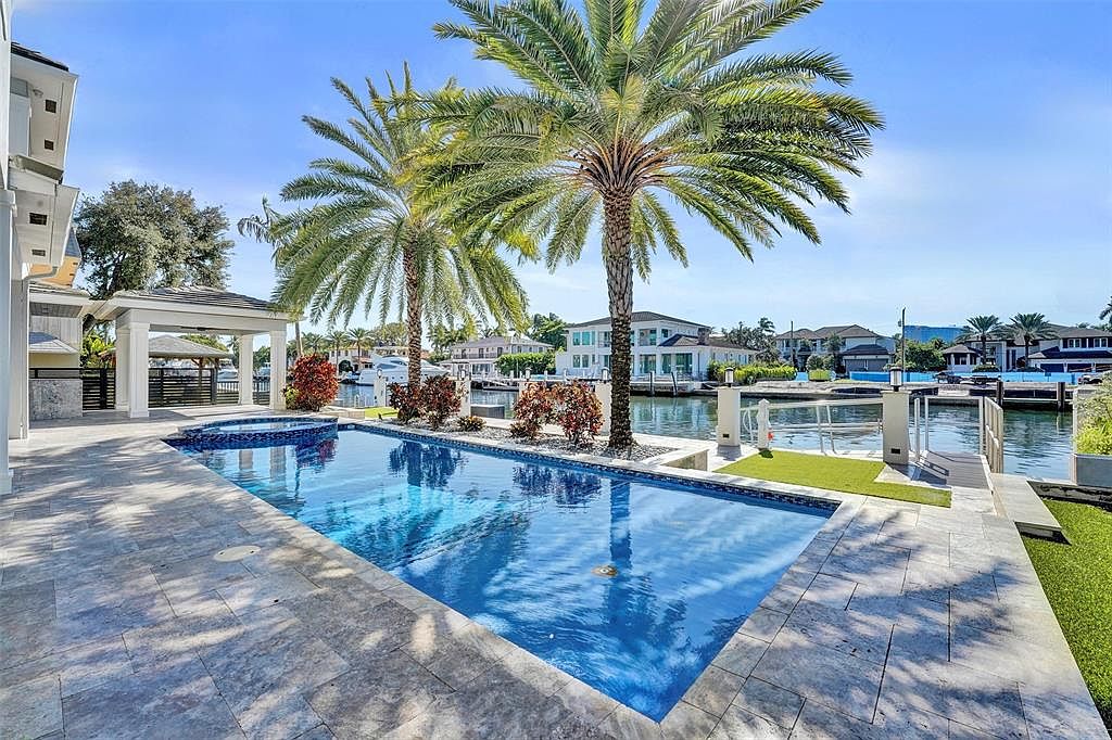 This image showcases a luxurious backyard featuring a rectangular pool with an attached spa, surrounded by travertine tile. Two palm trees stand prominently near the pool, adding a tropical feel. The property backs onto a waterway with a dock, suggesting waterfront access and upscale living.