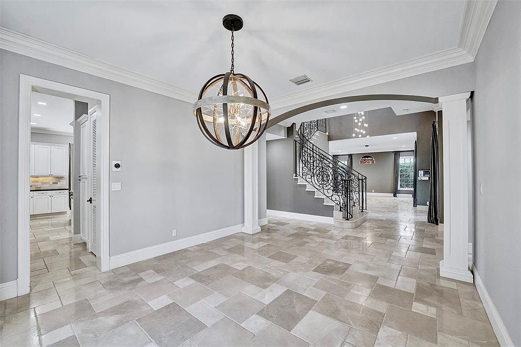 This interior shot showcases a dining room with a modern, yet classic design. The room features light gray walls, white trim, and a unique spherical chandelier. The flooring is a light-colored tile laid in a rectangular pattern, and the open layout leads to a staircase and other living areas, creating a sense of spaciousness and flow.