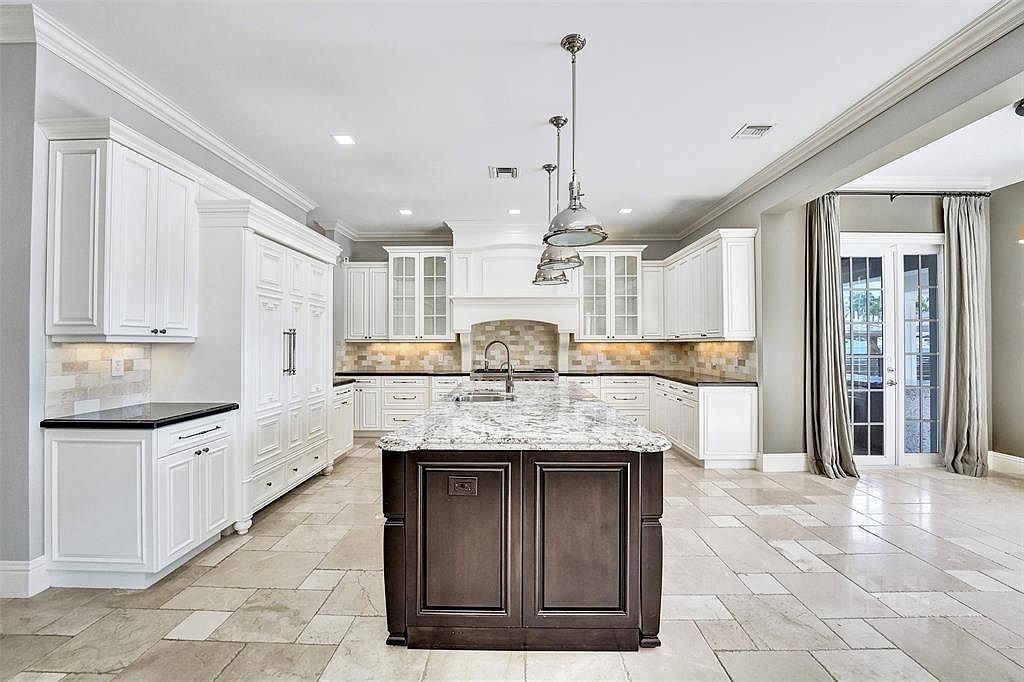This is a bright and spacious kitchen featuring white cabinetry, dark countertops, and a large center island with a marble countertop. The kitchen is well-lit with pendant lighting and recessed lights, and the flooring appears to be tile. The overall impression is one of luxury and functionality, perfect for a modern home.