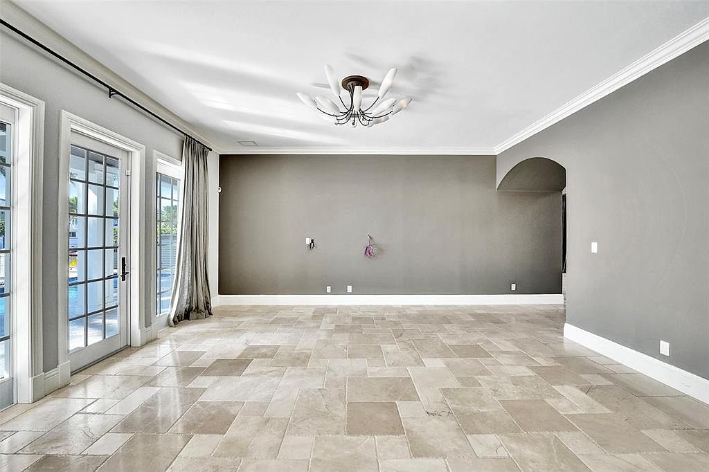 This is a spacious living room featuring neutral gray walls, a modern chandelier, and light-colored tile flooring. The room has large glass doors leading to an outdoor area, providing ample natural light. The overall impression is clean and contemporary, with a focus on open space.