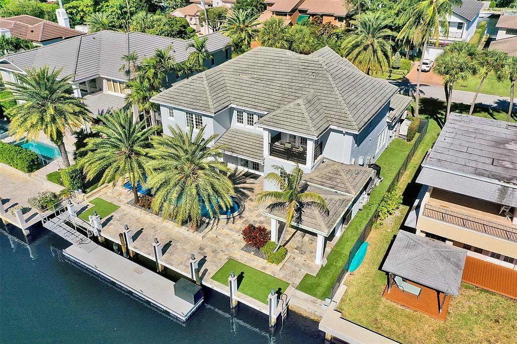 This aerial shot showcases a luxurious waterfront property with a large house featuring a gray tile roof, a private dock, and a well-manicured yard. The property includes a pool, a covered patio area, and mature palm trees, creating a tropical and upscale ambiance. The perspective is from above, providing a comprehensive view of the property's layout and its integration with the waterfront.