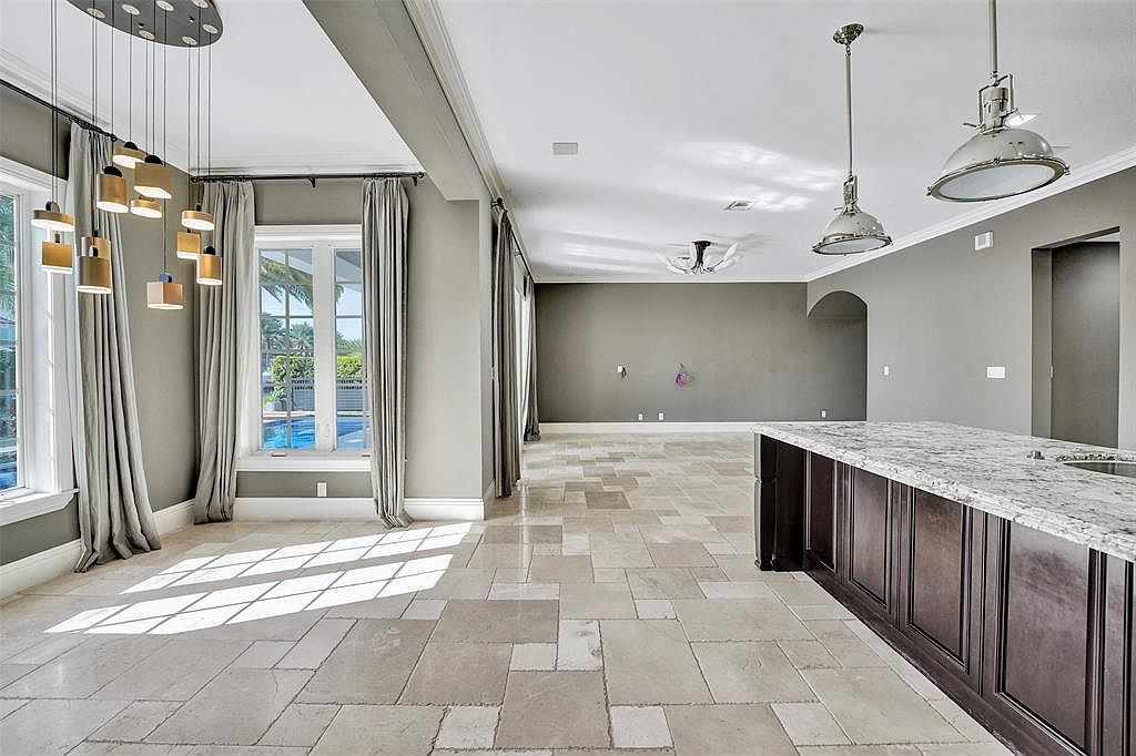This interior shot showcases a spacious living room with travertine flooring and neutral gray walls. Large windows with elegant drapery provide ample natural light and a view of the pool. Modern pendant lighting and industrial-style pendant lights above the kitchen island add a touch of sophistication to the space.