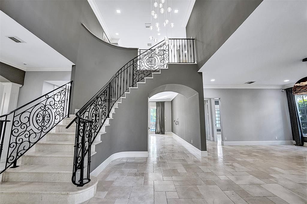 This interior shot showcases a grand foyer with a curved staircase featuring ornate black iron railings and light-colored stone steps. The walls are painted in a neutral gray tone, complemented by light tile flooring. An arched doorway leads to another room, and a modern chandelier hangs above the staircase, creating an elegant and spacious entrance.