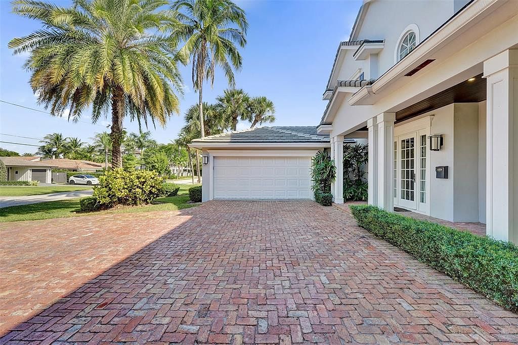 This is a front exterior view of a luxurious home featuring a brick driveway, well-manicured hedges, and palm trees. The house has a white garage door and a covered entryway with white columns and a set of double doors. The overall impression is one of elegance and curb appeal.