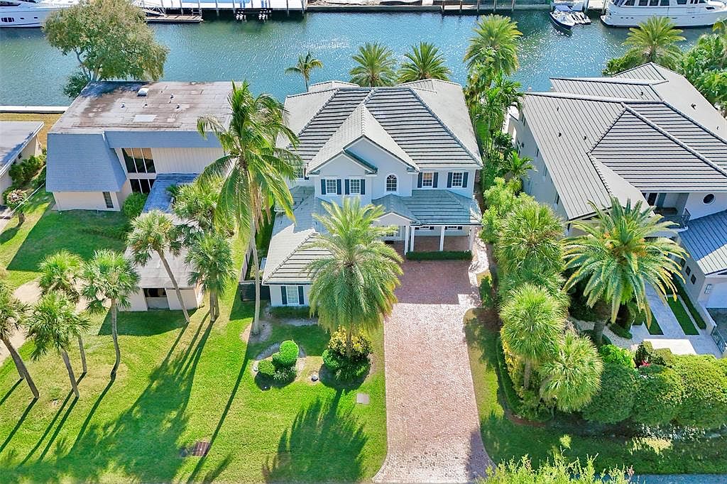 This aerial shot showcases a beautiful waterfront property with lush green lawns and mature palm trees. The house features a gray tiled roof, a brick driveway, and a well-maintained exterior. The property is situated on a canal, offering direct access to the water, enhancing its appeal as a desirable real estate option.