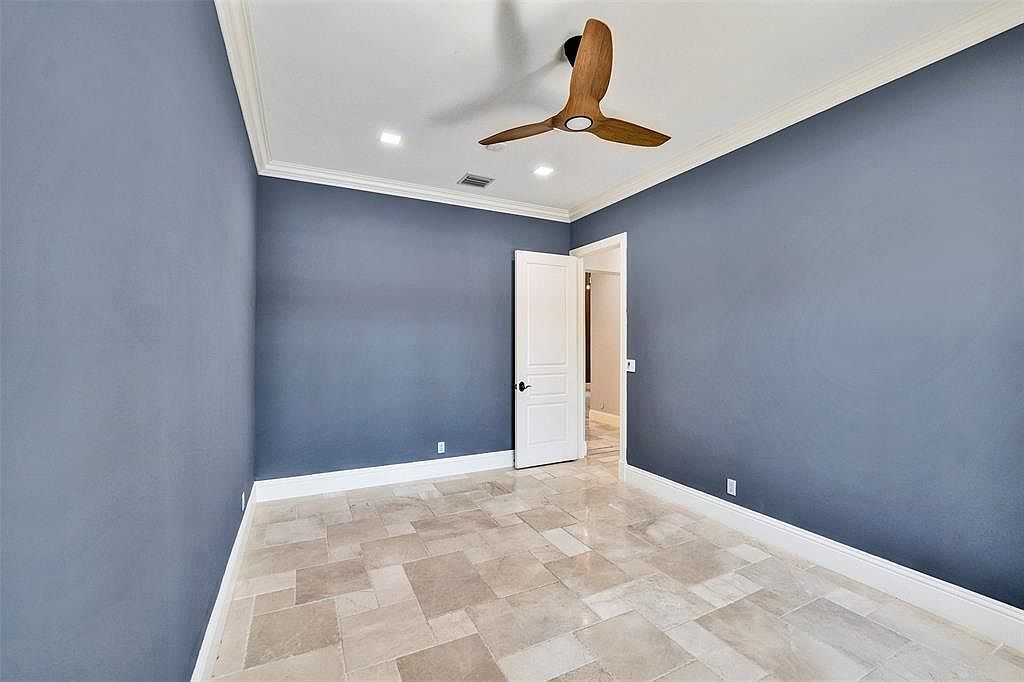 This is an interior shot of a bedroom featuring blue walls, a light-colored tile floor, and white trim. A wooden ceiling fan is mounted on the white ceiling. An open white door leads to another room, suggesting an en-suite bathroom or walk-in closet.