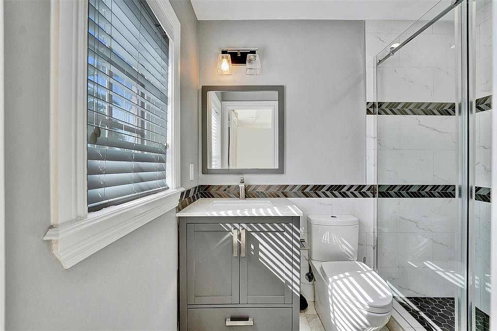 This is a well-lit bathroom featuring a gray vanity with a white countertop, a square mirror, and a modern light fixture. The shower is enclosed with glass and has decorative tile accents. The toilet is modern and the overall design is clean and contemporary.