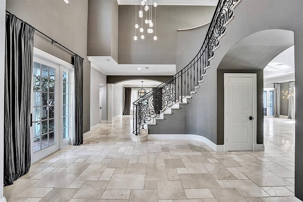 This grand foyer features a sweeping staircase with ornate iron railings, complemented by a high ceiling and elegant chandelier. The neutral color palette and travertine flooring create a sophisticated and inviting atmosphere. French doors to the left provide natural light and a view of the outdoors.