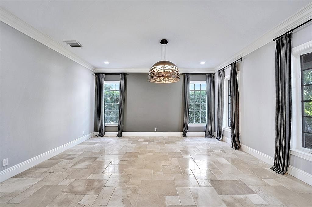 This is a spacious dining room featuring travertine tile flooring and light gray walls. The room is illuminated by a modern pendant light fixture and recessed lighting. Large windows with dark gray curtains provide natural light and a view of the outdoors, creating an elegant and inviting atmosphere.