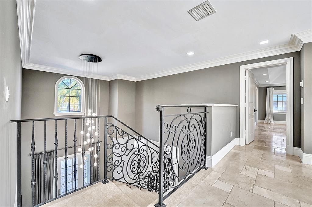 This interior shot showcases a well-lit hallway and staircase area. The space features elegant wrought iron railings, a modern chandelier, and a window providing natural light. The walls are painted in a neutral tone, complemented by white trim and light-colored tile flooring, creating a sophisticated and inviting atmosphere.