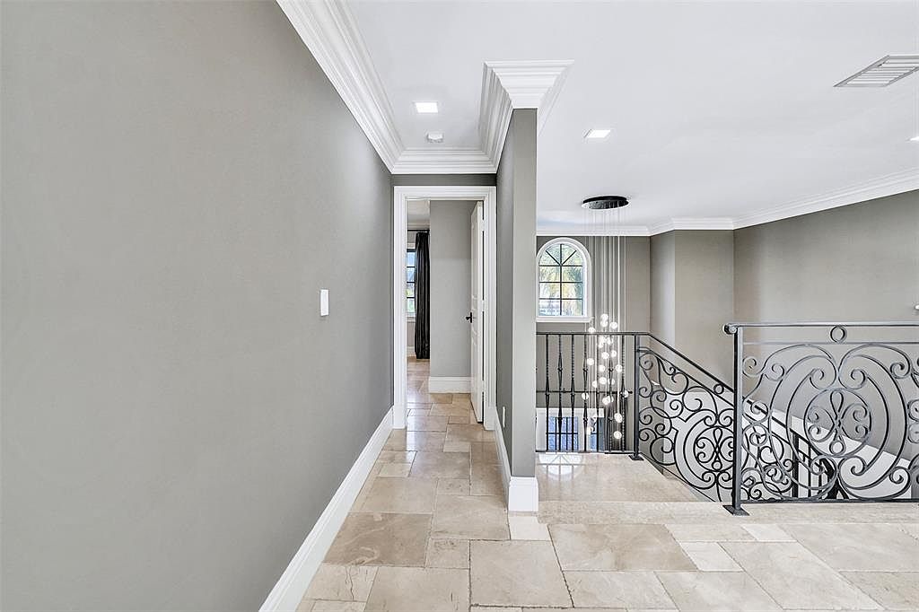This interior shot showcases a hallway with elegant architectural details, including crown molding and a decorative wrought iron railing overlooking a lower level. The flooring is tiled, and the walls are painted in a neutral gray tone, creating a sophisticated and inviting atmosphere. A doorway leads to another room, hinting at the home's spacious layout.