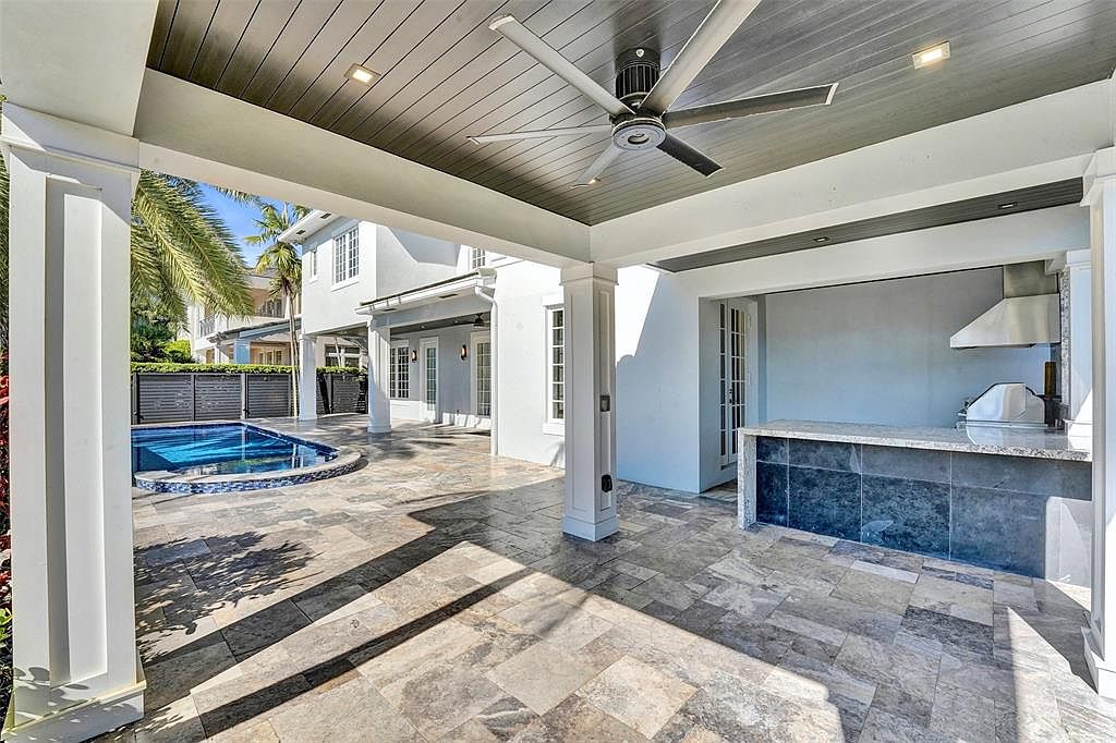 This image showcases a luxurious outdoor patio area, featuring a built-in outdoor kitchen with a stainless steel grill and countertop. The patio is covered with a ceiling fan and recessed lighting, and the flooring is composed of large, patterned stone tiles. A swimming pool is visible in the background, adding to the upscale ambiance of the outdoor living space.