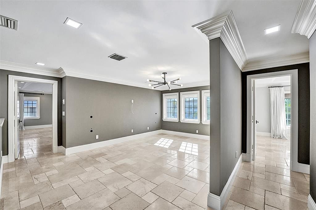This is an interior shot of a bedroom featuring neutral gray walls, white trim, and travertine tile flooring. The room has multiple windows providing natural light and a modern chandelier. The space appears spacious and well-lit, suitable for a guest bedroom or secondary bedroom.