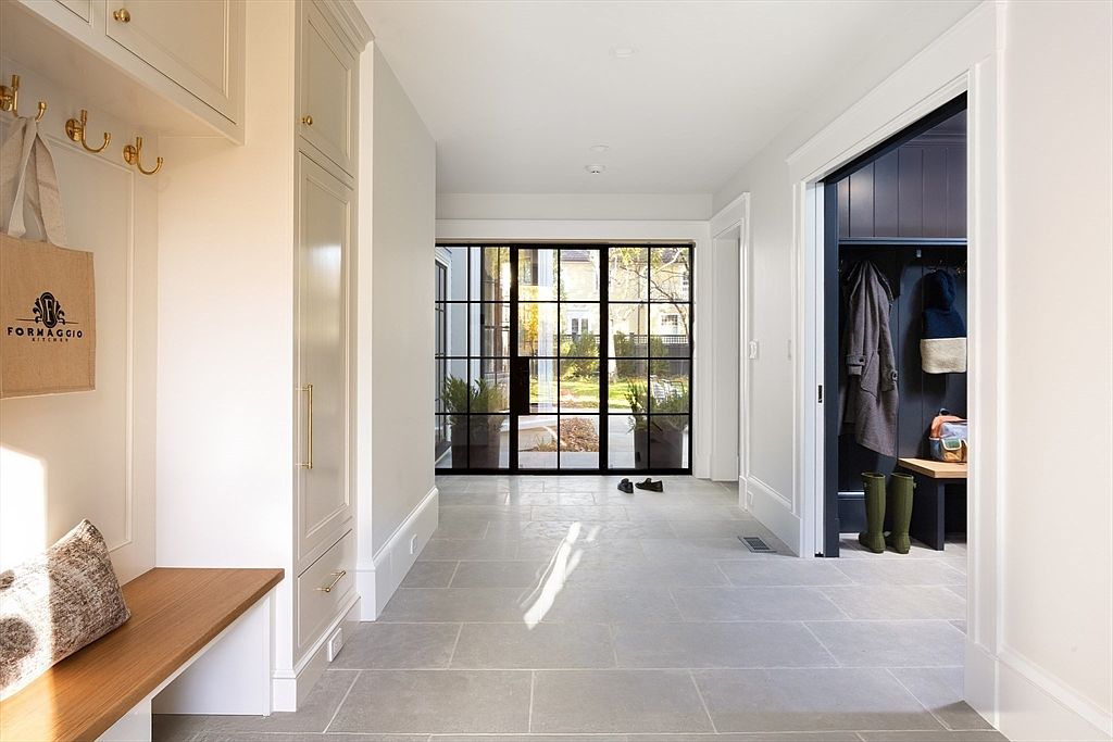 This bright and airy entryway features elegant light-colored stone flooring and custom built-in cabinetry with brass hardware. A striking black-framed glass door at the end of the hall provides a clear view of the outdoor landscape, while a separate mudroom area with dark cabinetry adds functional contrast. The space feels sophisticated and welcoming, blending modern architectural lines with practical storage solutions.