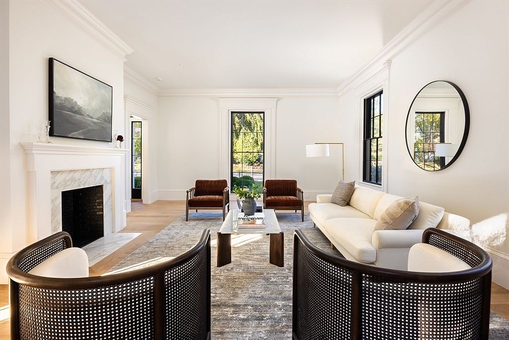 This elegant living room features a sophisticated, neutral color palette with a central fireplace framed by a marble surround and a large landscape painting. The space is furnished with a plush white sofa, two rich brown velvet armchairs, and two curved, cane-backed accent chairs in the foreground, all arranged on a textured area rug. Large windows with black frames allow natural light to flood the room, creating an airy and refined atmosphere perfect for high-end residential living.