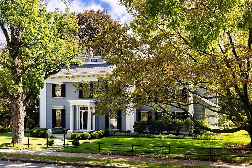 This classic, two-story colonial-style home features elegant grey siding, crisp white trim, and symmetrical black shutters, exuding a timeless and stately curb appeal. The front entrance is highlighted by a charming portico, while a manicured lawn and mature, leafy trees frame the property, creating a serene and inviting atmosphere. The perspective is a straight-on, eye-level shot that captures the full architectural beauty of the home from the street.