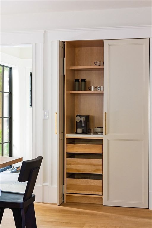 This image showcases a custom-built, floor-to-ceiling pantry cabinet with a hidden coffee station, featuring light wood shelving and drawers contrasted against elegant cream-colored doors with brass hardware. The perspective is a medium shot from the side, highlighting the functional design of the pull-out drawers and the integrated appliance nook. The clean lines and warm wood tones create a sophisticated, organized, and modern aesthetic suitable for a high-end residential listing.