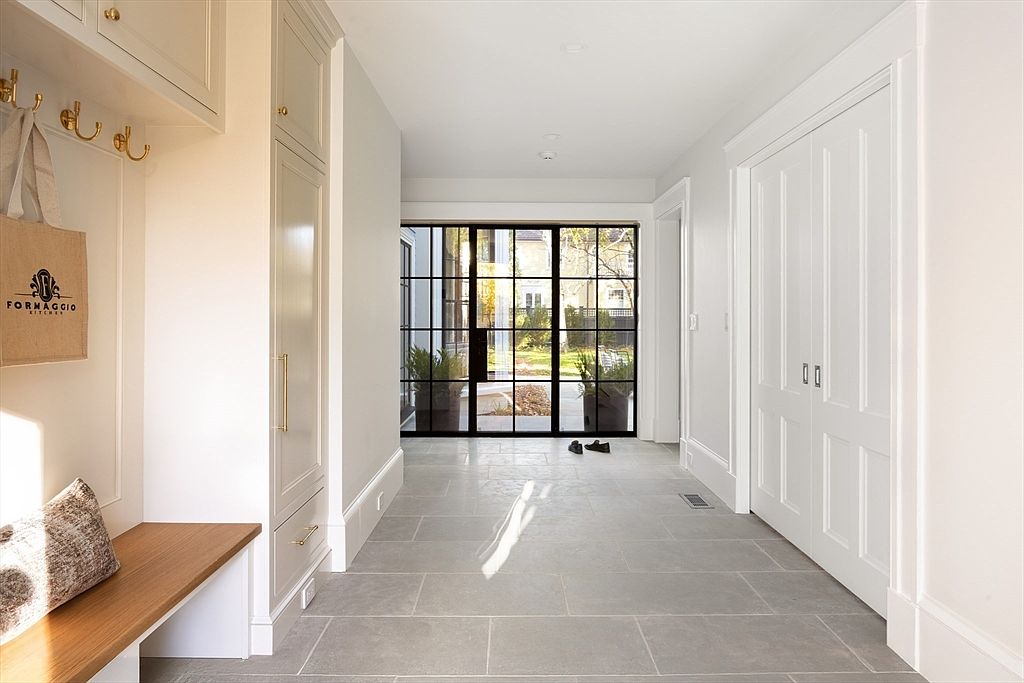 This elegant entryway or mudroom features a clean, transitional design with light gray stone flooring and crisp white walls. To the left, a built-in bench with storage and coat hooks provides functional organization, while a large, black-framed glass door at the end of the hall offers a bright, inviting view of the outdoor garden. The space is balanced by a set of white double doors on the right, creating a symmetrical and sophisticated transition area.