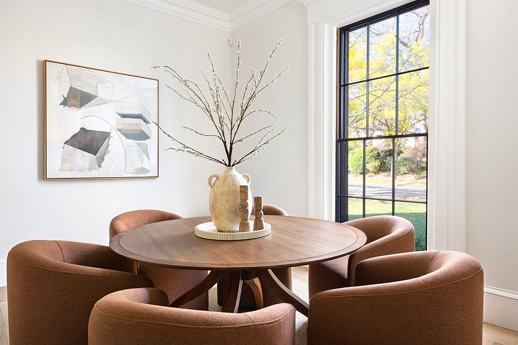 This elegant dining area features a round wooden table surrounded by four plush, rust-colored upholstered chairs, creating a warm and inviting atmosphere. A large, textured ceramic vase filled with delicate branches serves as a sophisticated centerpiece, complemented by a piece of abstract wall art. The space is bathed in natural light from a large, black-framed window, highlighting the clean lines and modern aesthetic of the room.