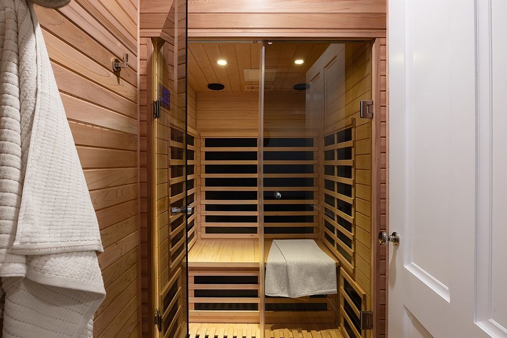 This image showcases a compact, modern home sauna constructed with light-toned cedar wood paneling and a glass door. The interior features slatted wooden benches and integrated heating panels, creating a clean, spa-like atmosphere. A white towel hangs on the exterior wall, emphasizing the wellness-focused nature of this dedicated relaxation space.