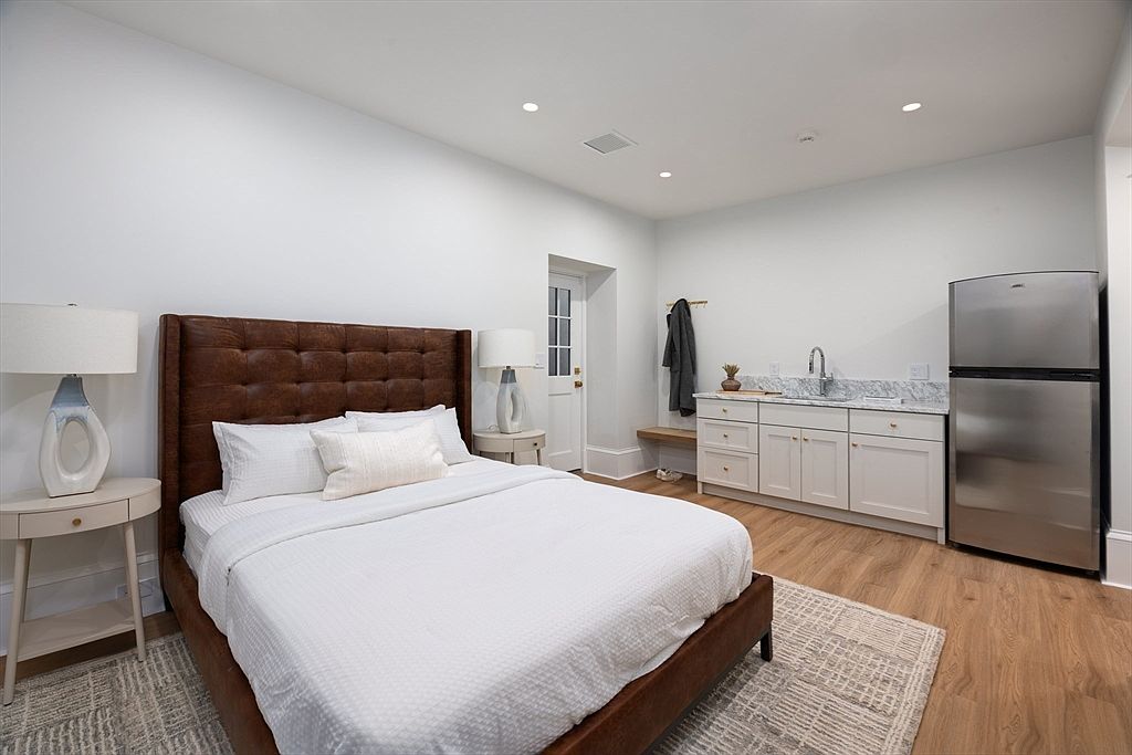 This well-appointed bedroom features a rich, tufted brown leather headboard as the focal point, complemented by a neutral color palette and light wood flooring. The room includes a convenient kitchenette area with white cabinetry, a marble countertop, and a stainless steel refrigerator, making it ideal for guests. The perspective is a wide-angle shot that captures the entire functional layout, emphasizing the blend of comfort and utility.