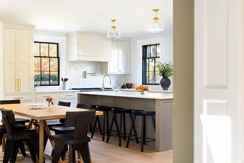 This bright and airy kitchen features a sophisticated blend of cream-colored perimeter cabinetry and a contrasting dark gray central island topped with white marble countertops. The space is anchored by a wooden dining table in the foreground, complemented by black modern chairs and stools, while elegant glass pendant lights hang above the island. Large windows with black frames provide ample natural light, creating a clean, high-end, and inviting atmosphere perfect for both cooking and entertaining.