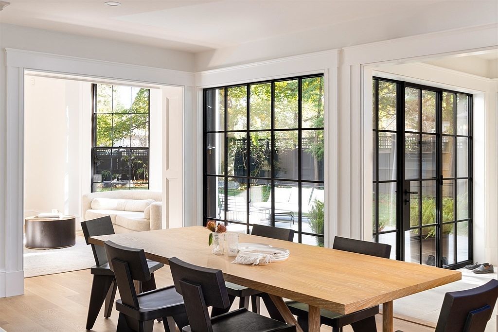 This elegant dining room features a large, light-toned wooden table surrounded by modern black chairs, creating a sophisticated contrast. The space is defined by expansive, floor-to-ceiling black-framed windows that flood the room with natural light and offer a seamless view of the outdoor patio and garden. The clean lines, neutral color palette, and architectural detailing evoke a refined, contemporary aesthetic perfect for upscale living.