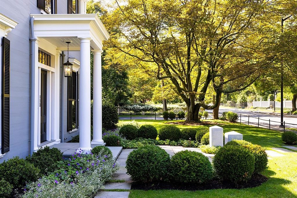 This elegant front entryway features a classic portico supported by white columns, leading to a dark-painted door with a traditional lantern fixture. The home is surrounded by meticulously manicured boxwood shrubs and vibrant purple flowers, set against a backdrop of a lush, sun-drenched lawn and mature trees. The perspective captures a welcoming, high-end residential curb appeal with a clear view of the stone walkway and the surrounding neighborhood landscape.