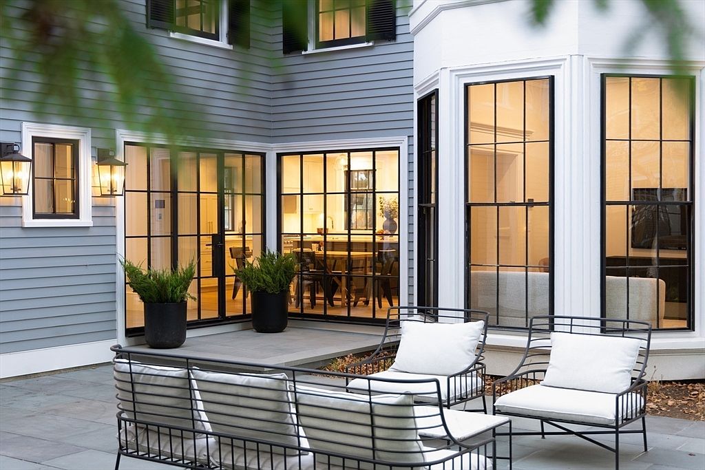 This elegant outdoor patio space features modern black-framed glass doors and windows that seamlessly connect the indoor living area to the stone-paved terrace. The scene is furnished with contemporary black metal seating topped with plush white cushions, creating a sophisticated and inviting atmosphere. The gray siding and warm interior lighting visible through the glass provide a cozy, high-end residential aesthetic.